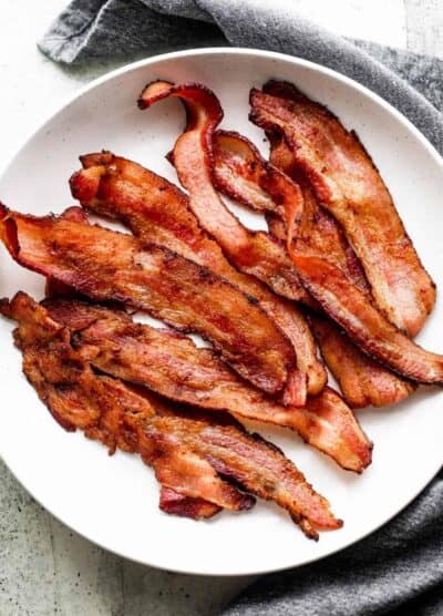 strips of air fryer bacon arranged on a white plate, with a gray tea towel placed underneath the plate.