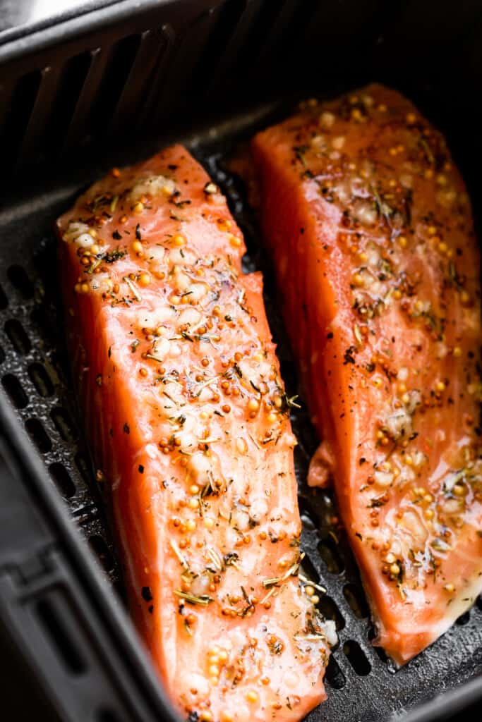 two raw salmon fillets topped with mustard dressing and set inside a black air fryer basket.