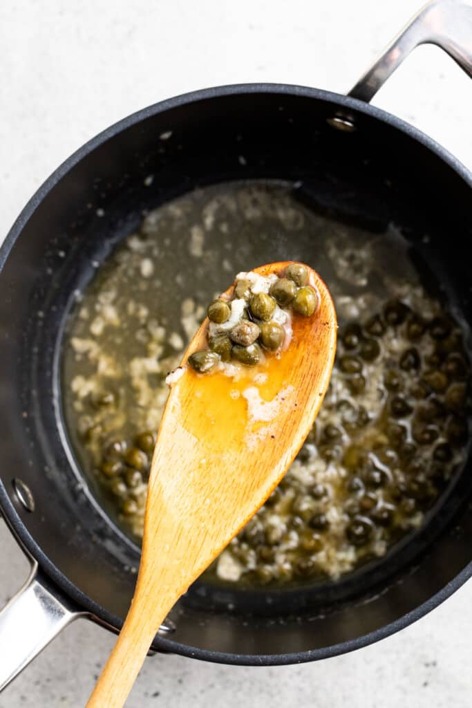 A wooden spoon with lemon caper sauce hovering over a saucepan of lemon caper sauce.