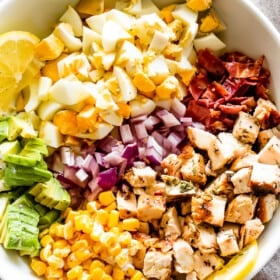 overhead shot of a white salad bowl with chopped hard boiled eggs, sliced avocados, diced onions, diced cooked chicken, crumbled bacon, and corn kernels.