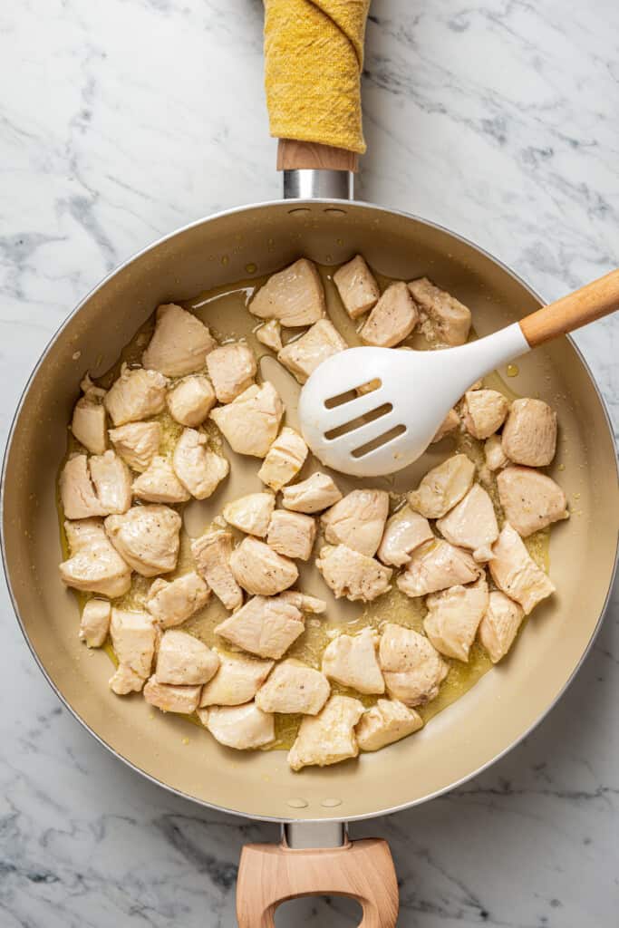 A skillet with chunks of chicken being stirred by a slotted spoon