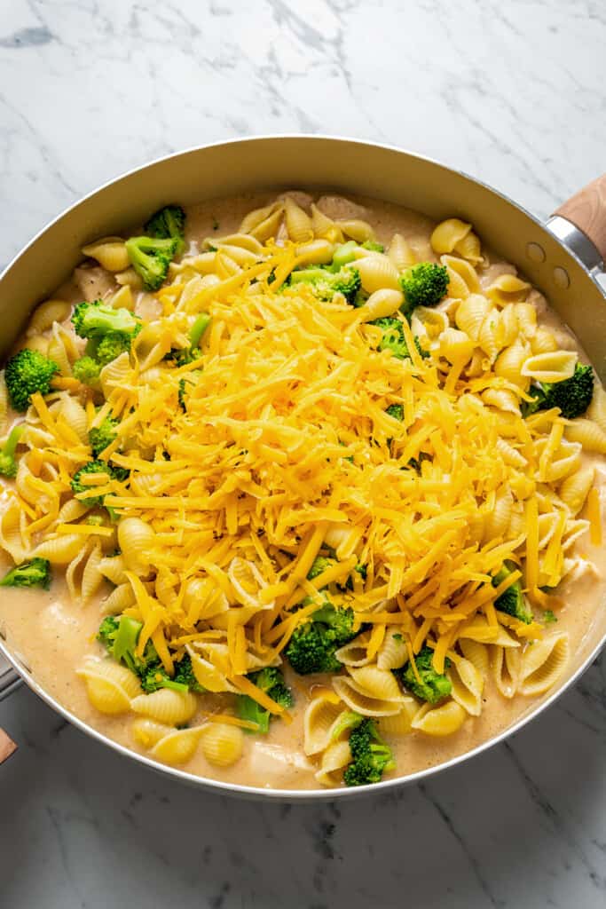A pile of broccoli and shredded cheese in a skillet on top of pasta, chicken, and sauce
