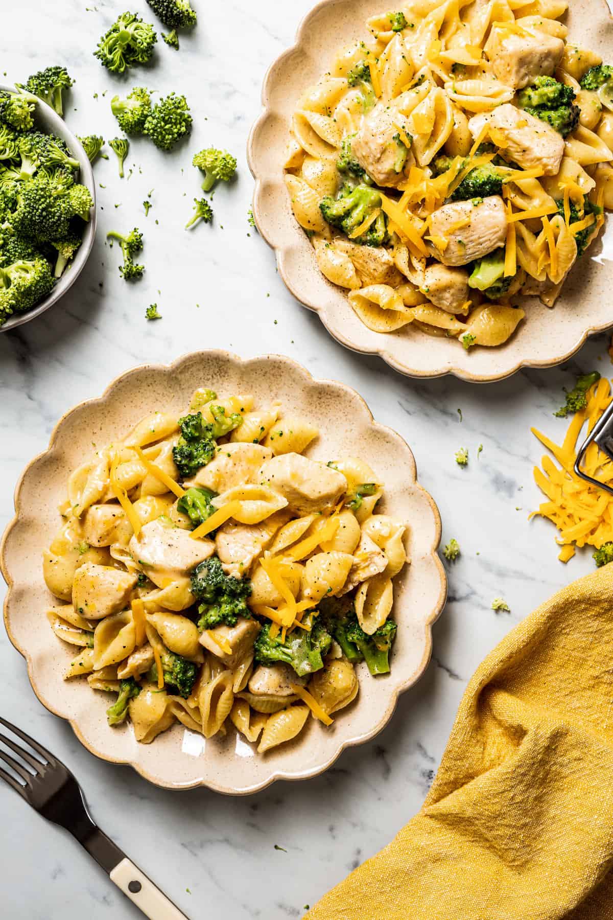 Overhead view of two plates of broccoli and chicken pasta, next to a bowl of broccoli, a fork, and a kitchen towel.