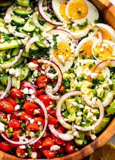 close up of sliced red onions, halved hard boiled eggs, chopped avocado, sliced cherry tomatoes, and sliced cucumbers topped with crumbled feta cheese
