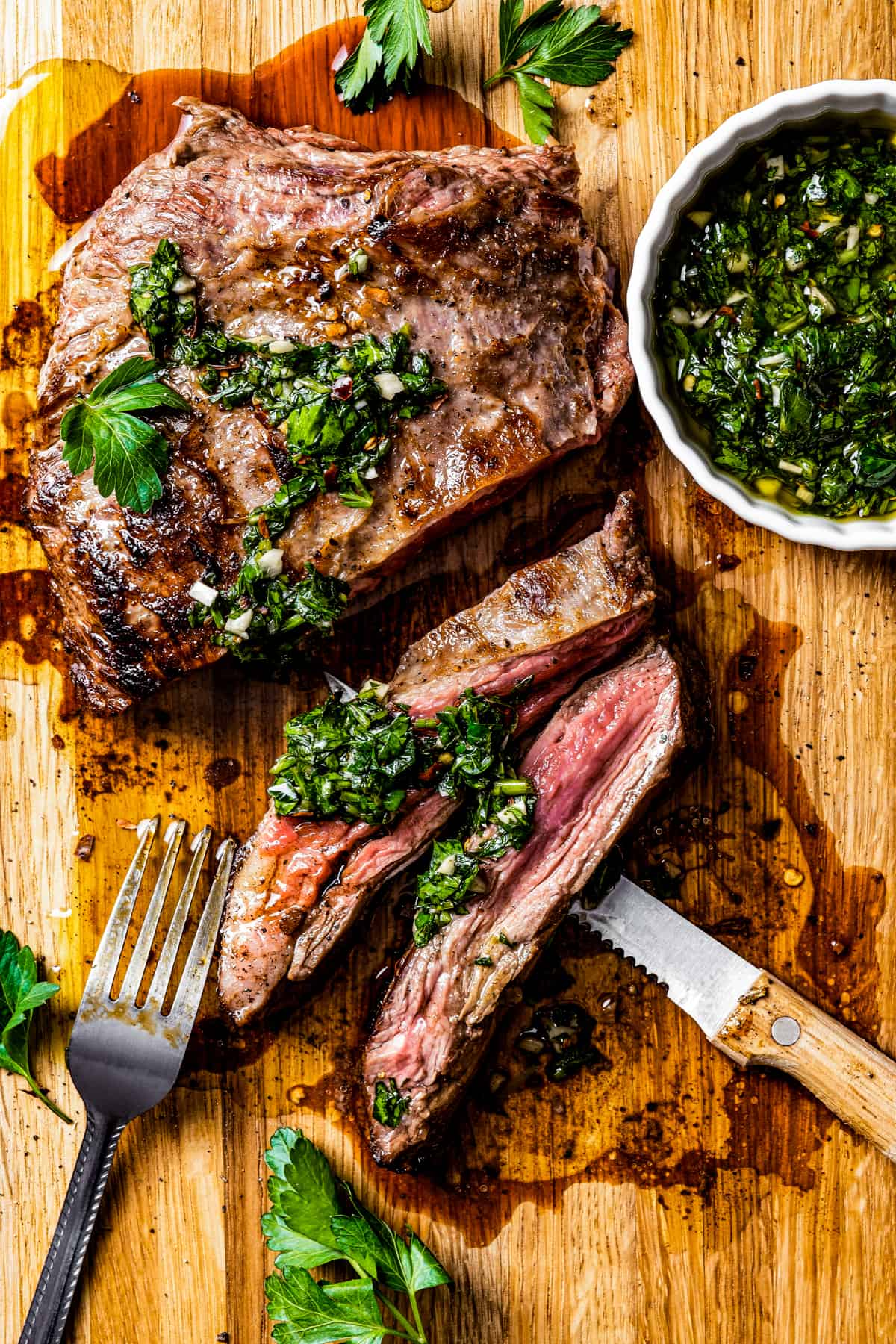 Churrasco steak on a cutting board with a knife and fork and a bowl of chimichurri.