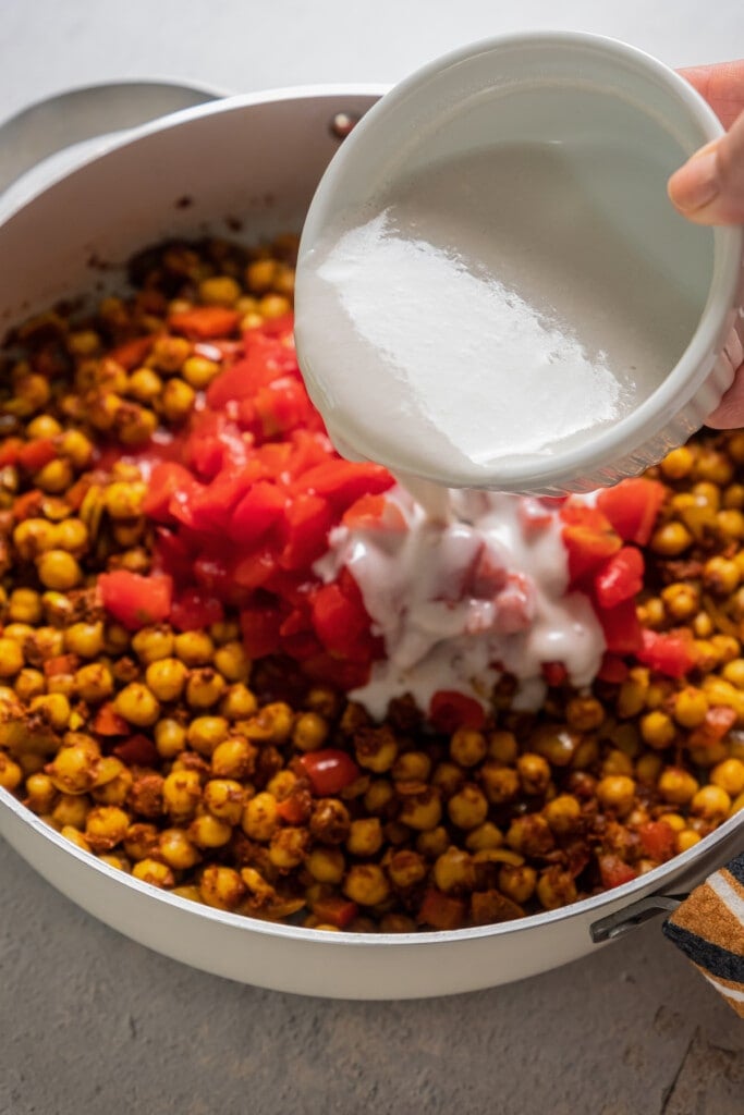 Adding tomatoes and coconut milk to toasted chickpeas in a saucepan.