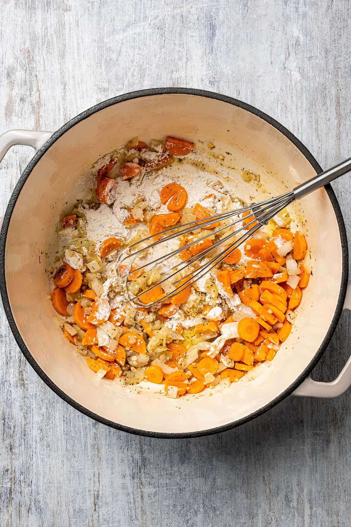 A roux made of flour and butter is whisked into sautéed onion, garlic, and carrots in a pot.