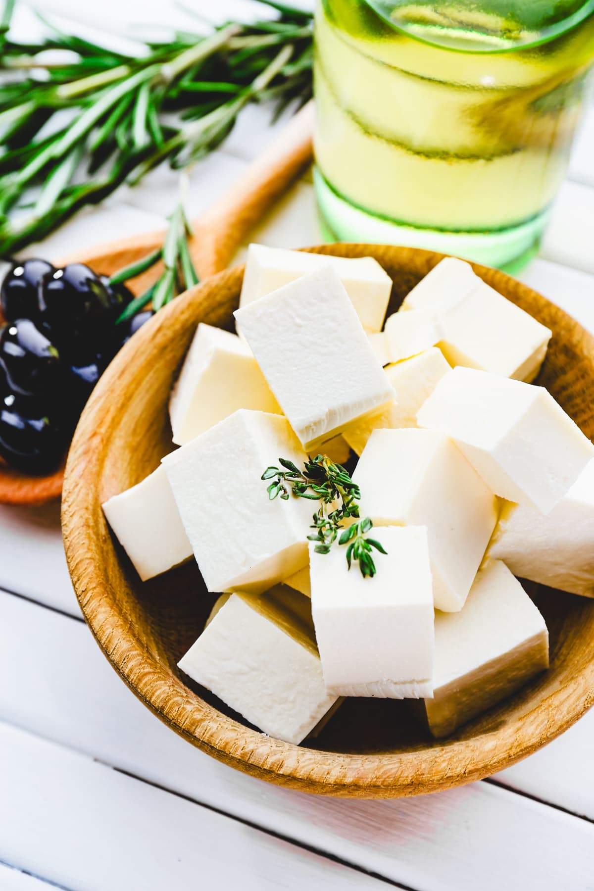 Feta cheese cubes in a brown wooden bowl, with a rosemary sprig, olive oil, and olives set behind the bowl.