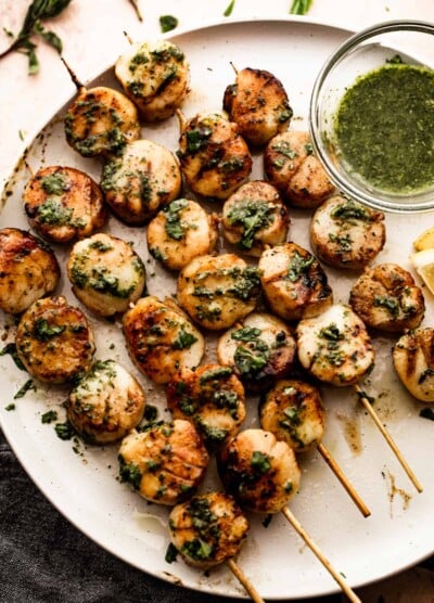 grilled scallops threaded onto five skewers and arranged on a white plate with a small bowl of basil and garlic sauce placed next to them.