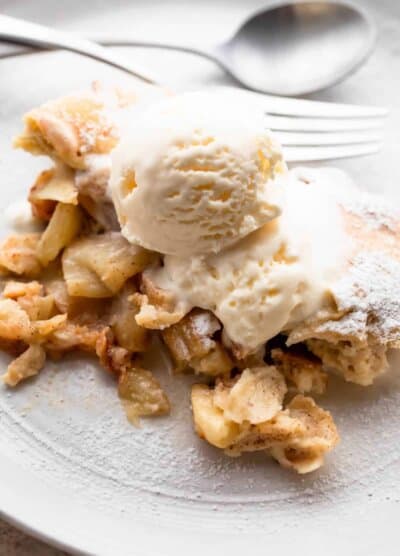 healthy apple cobbler served on a white plate, topped with a scoop of vanilla ice cream, and a spoon and fork set near the dessert.