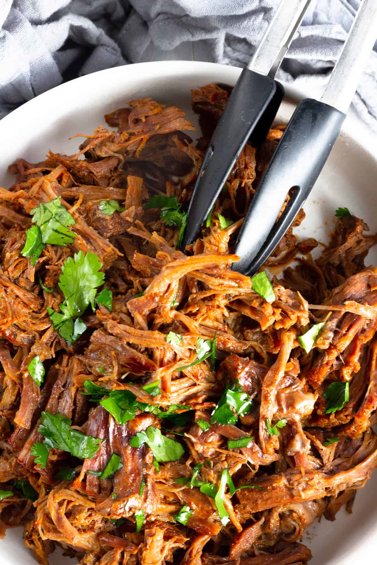 A bowl with beef barbacoa and tongs lifting up the beef.