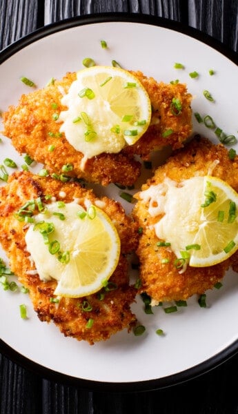 overhead shot of three breaded chicken breasts, also known as chicken milanese, arranged on a plate and topped with lemon slices.