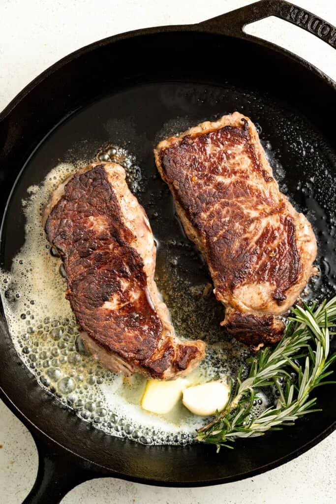 Adding butter, garlic, and rosemary to a pan with seared steak.