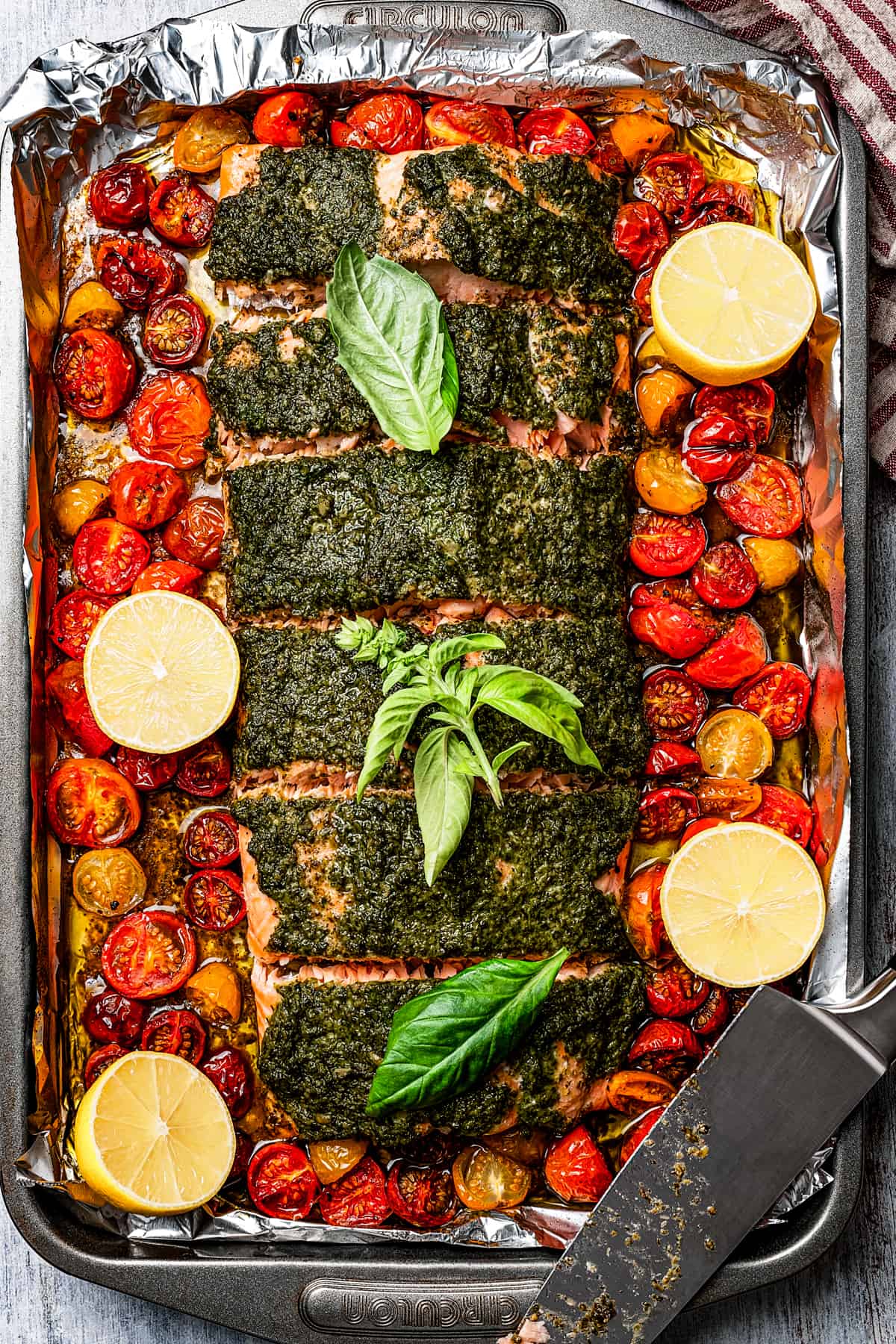 A tray of baked salmon with pesto, cherry tomatoes, and lemons.