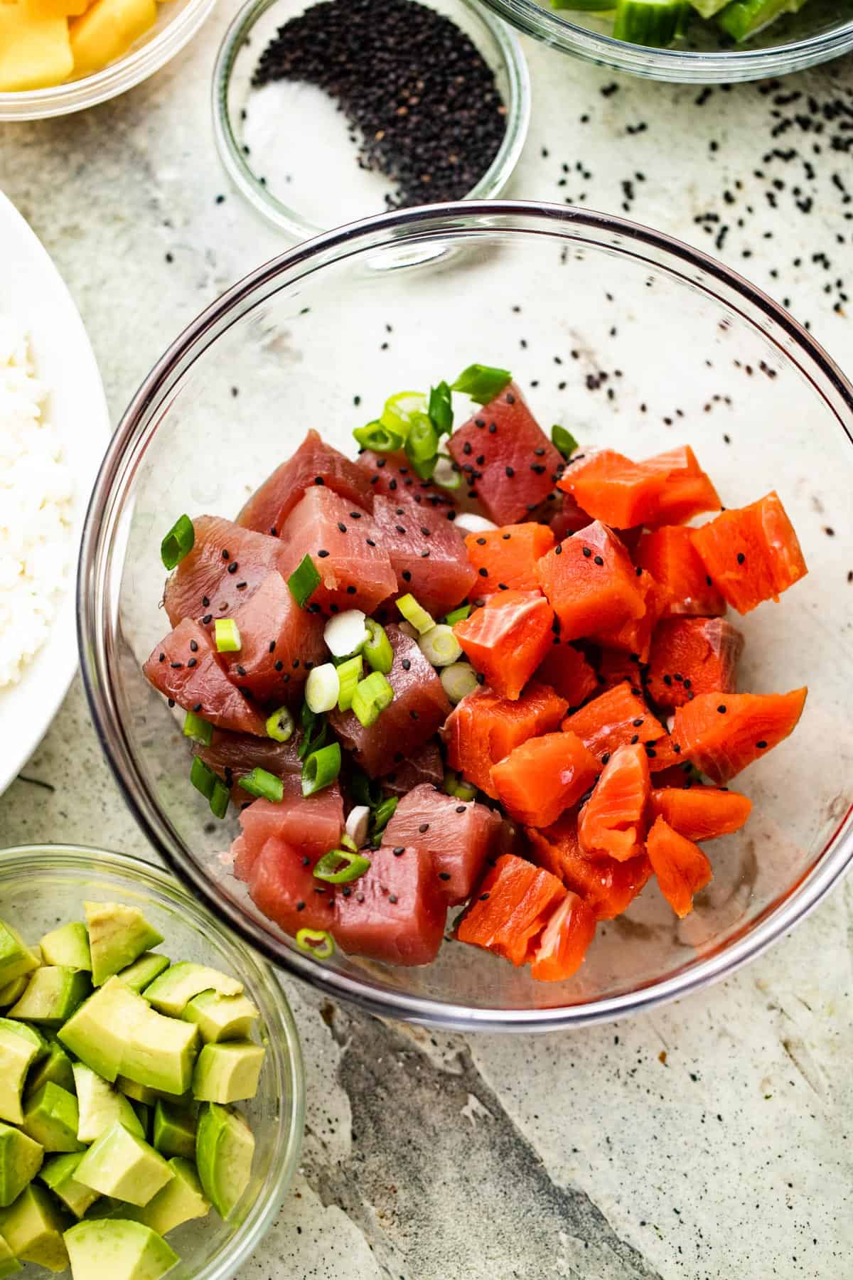glass bowl with raw pieces of tuna, raw pieces of salmon, with a bowl of diced avocado to the left, mango chunks above, and black poppy seeds set above.