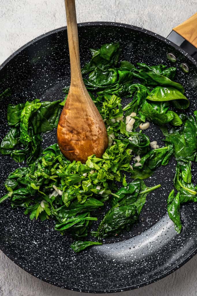 A wooden spoon stirring wilted spinach with garlic in a black skillet.