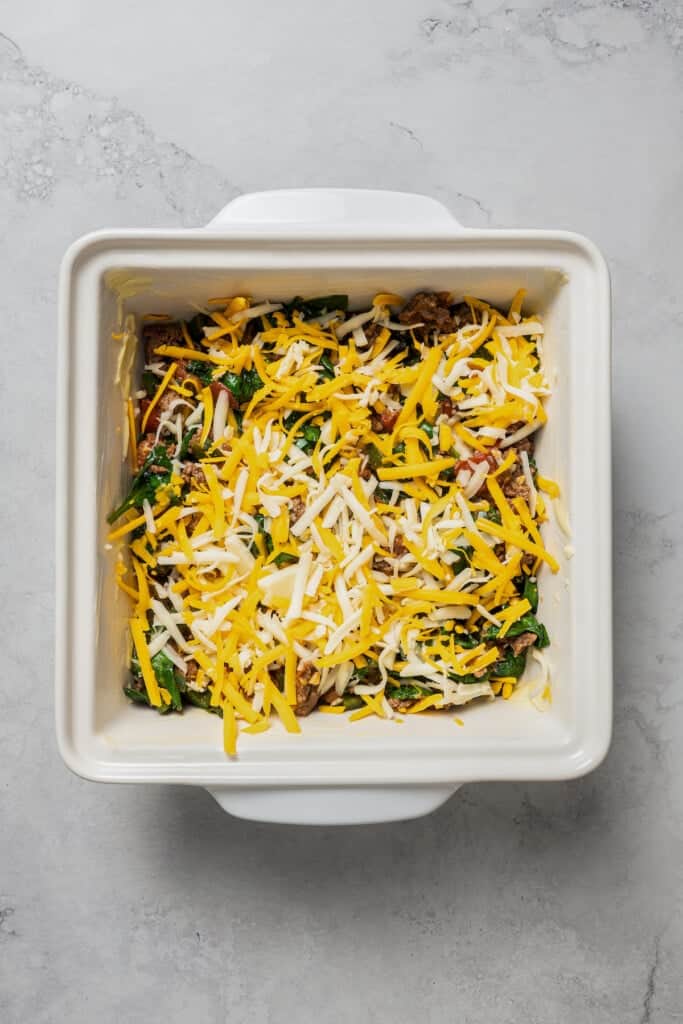 A partially assembled taco casserole in a square baking dish, topped with shredded cheese.