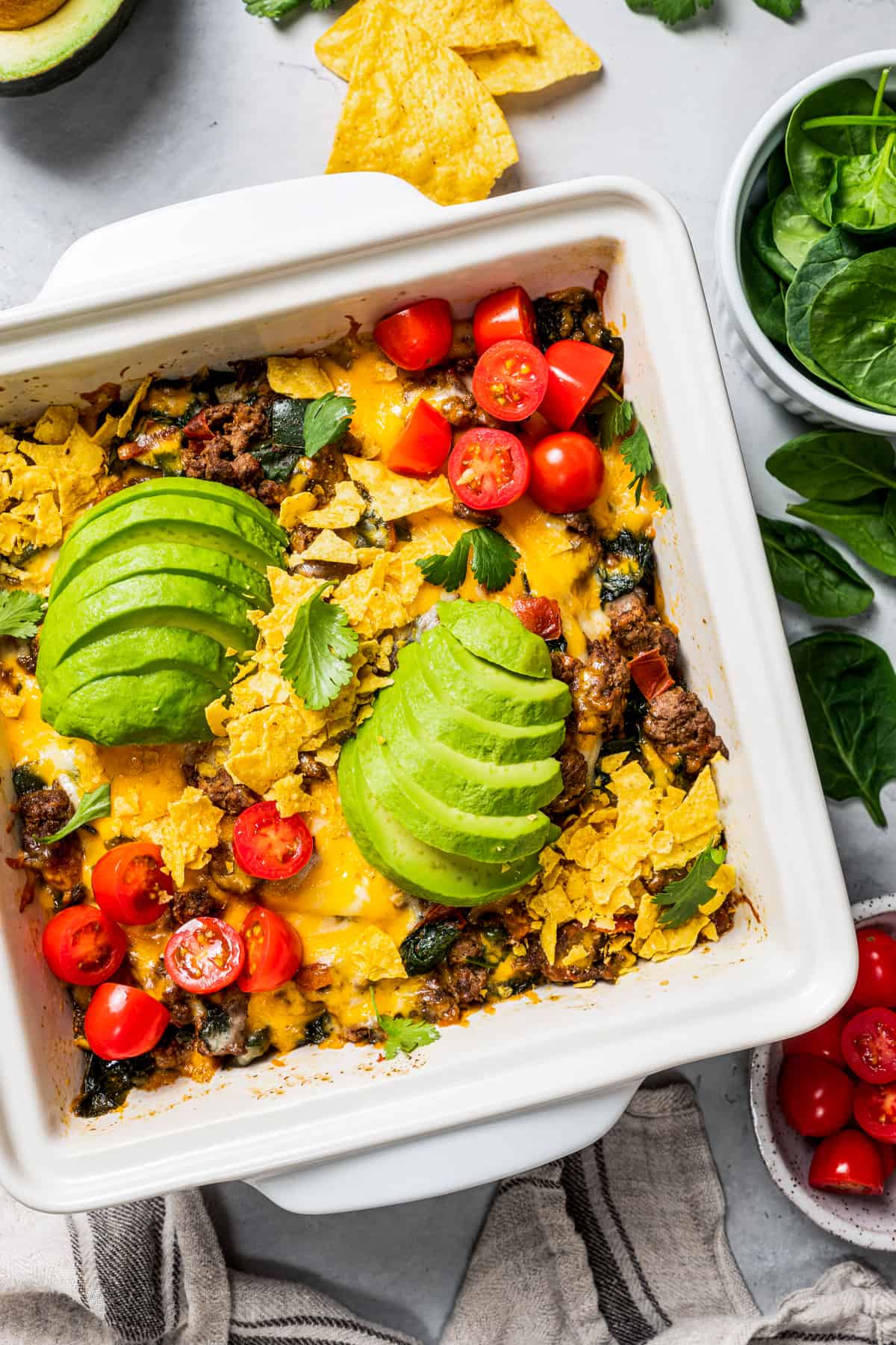 Overhead view of a baked taco casserole in a square baking dish topped with sliced avocados, cherry tomatoes, and fresh cilantro.