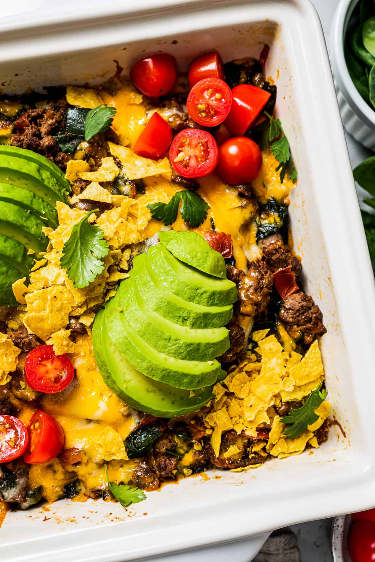 Close up overhead view of a baked taco casserole in a square baking dish topped with sliced avocados, cherry tomatoes, and fresh cilantro.