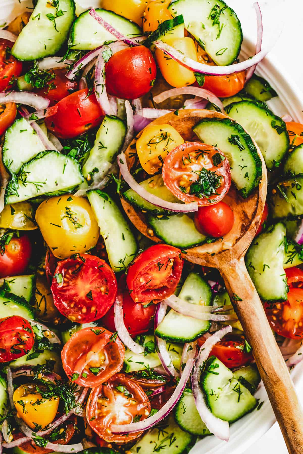 Tomato and cucumber salad in a serving bowl with a wooden spoon scooping out of the salad.