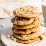 Six white chocolate chip cookies stacked on top of each other on a plate, surrounded by white chocolate chips